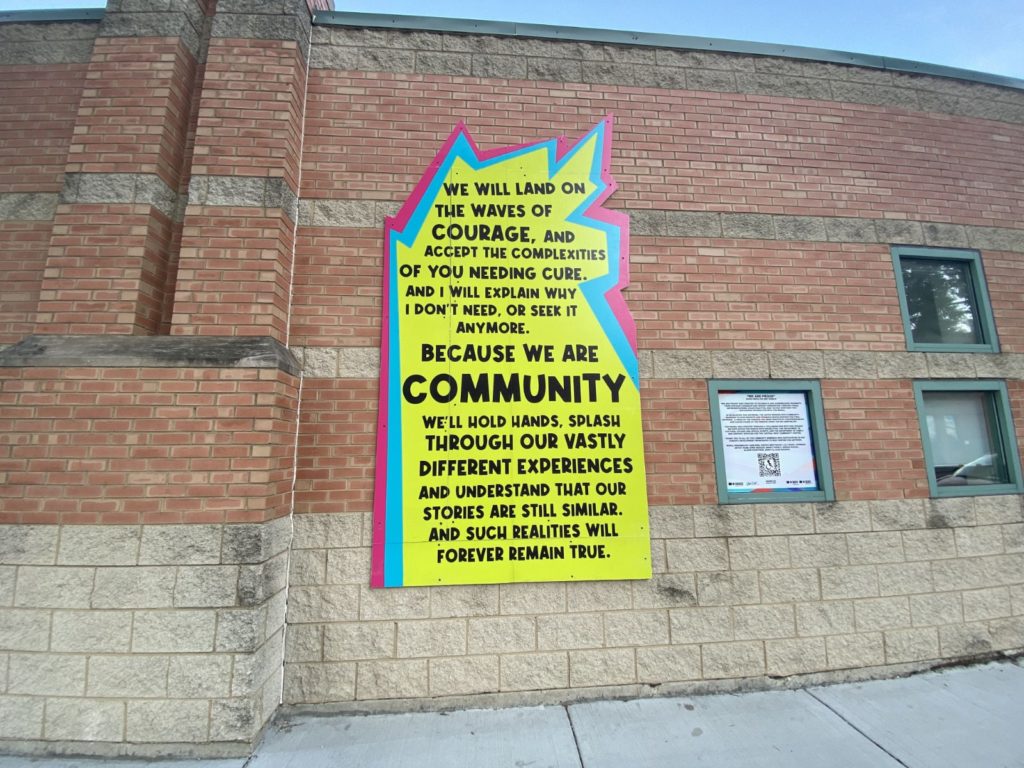 The first section of the mural. It is a bookend before the images start. It is an abstract shape with a clean square shape on the bottom but a jagged and pointy shape at the top. It is mostly a bright yellow-green color, with light blue and bright pink edges bordering it. Inside of the shape are black bubble letters. The text says, “We will land on the waves of courage, and accept the complexities of you needing cure. And I will explain why I don’t need, or seek it anymore. Because we are community. We’ll hold hands, splash through our vastly different experiences, and understand that our stories are still similar. And such realities will forever remain true.” The biggest words on the sign are the phrase, “Because we are community.”