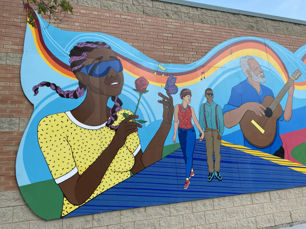 One section of the first stretch of the mural along the side of MOPD Walls. A girl with black skin wears glasses for the blind and holds a rose in one hand, and a butterfly sits on the pointer finger of her other hand. She is smiling and wearing a yellow shirt. A couple holds hands and walks down a blue path. They do not have obvious or visible disabilities. The woman, on the left, has light brown skin, wears a tank top, blue jeans, red shoes, and wears her hair wrapped on top of her head. The man, on the right, has dark skin, wears sunglasses, a blue button down shirt, suspenders, khaki pants, and blue shoes. A man with blue eyes and gray hair plays the guitar, and lines depicting the movement of the music flow throughout the scene. These lines of sound are yellow, orange, and burgundy.