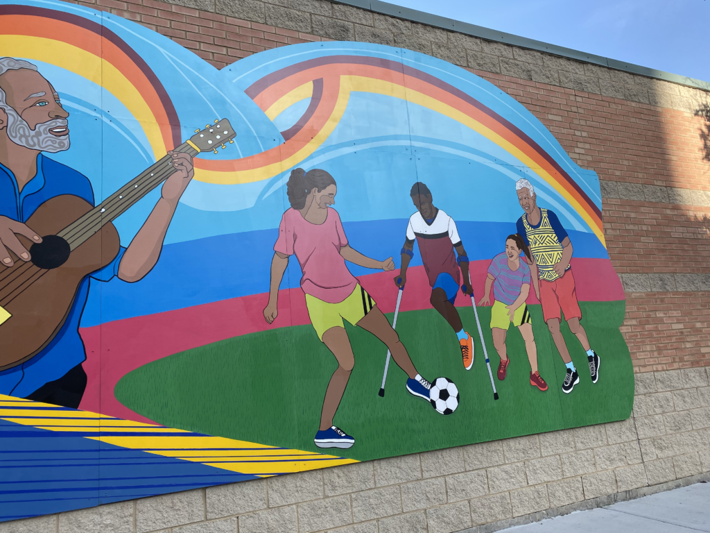 Another section of the first stretch of the mural along the side of MOPD Walls. A group of young people, including a person who only has one leg using crutches and an older man, plays soccer together on green grass. THere is a line of pink on the horizon, but mostly light blue to depict the sky. The person using crutches has black skin and black hair, and wears a shirt that is white at the top, has a black horizontal stripe through the chest, and is maroon at the bottom. He wears blue shorts, light blue socks, and orange shoes. To his left is a young woman with tan skin, brown hair tied in a ponytail, a pink shirt, bright green short switch two diagonal black lines through her left leg, and blue and white sneakers. She is kicking the soccer ball. To the right of the person using crutches is another young woman. Her skin is lighter, and she wears her brown hair also in a ponytail. Her shirt is purple with blue horizontal stripes, and she has the same shorts as the girl kicking the soccer ball. Her sneakers are two shades of light and dark red. To her right is an older man with tan skin. He has white, curly hair and wears a blue t-shirt with a yellow sweater vest over it. The vest has a triangular pattern on it. He also wears red shorts, light blue socks, and black and white sneakers. They are all smiling as they play soccer.