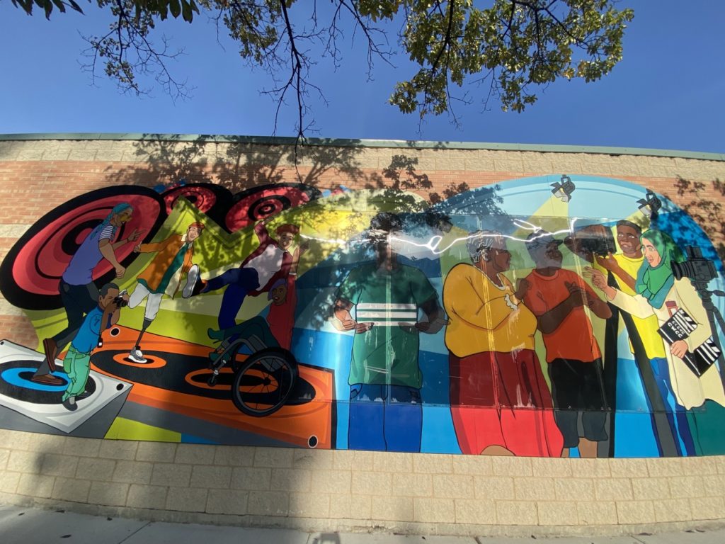 This is the full section of the second half of the mural as described in the previous two images, including the friends dancing, the boy smiling, and the group of people on the film set. Some of the image is covered by shadows of trees next to the building.