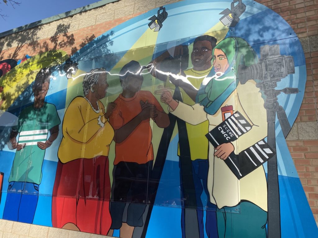 This is the scene on the right half of the section of the mural with the friends dancing. On the far left, behind a blue background, is a young boy with black skin and black hair. He wears a blue-green shirt with three white horizontal stripes in the center, and blue jeans. He is smiling, and his arms are bent at the elbows with his hands facing inward, palms up. His hands are moving. He is possibly using sign language to communicate. To the right of him is a group of people working on a film shoot. The first person is an older woman turned so that we can see her right side. She has a larger body than the other people in the scene. She has black skin, short black hair with lots of gray streaks, a yellow long sleeved shirt with a white collar and white sleeves, and a long red skirt. She has several piercings in her right ear, and wears a hearing aid. She is using her right hand to say “yes” in American sign language. Next to her is a younger boy. He has black skin, a blue baseball cap, an orange t-shirt, and navy blue pants. He is smiling and listening and is holding his hands together, in front of his chest. To his right is a taller man with black skin, short black hair, a light green t-shirt, and blue jeans. He is operating a camera and using his hands to hold it while it sits on a tripod. To his right is a muslim woman with green eyes. She wears a green and blue hijab, a cream colored tunic, and teal pants. She also has an orange and red badge on the top of her tunic, and is holding a film slate in her left arm. With her right arm, she is moving her hand. She is pointing her finger and moving it back and forth, perhaps using sign language. To her right is another film camera on a tripod. Above the scene are stage lights shining down on them, presumably attached to a ceiling.