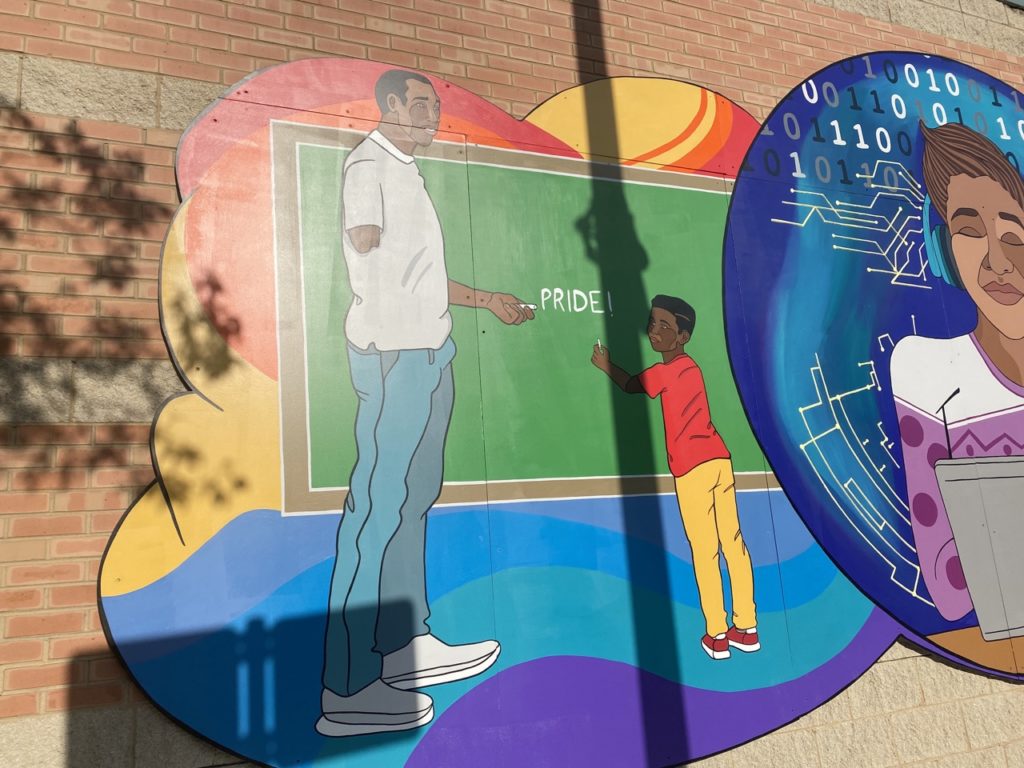 This is the leftmost side of the third scene of the mural. There is a rounded, rainbow swirling background behind the scene. The scene predominantly features a green chalkboard with a brown border. Written in the center of the chalkboard is the word PRIDE! In front of the chalkboard stands a tall man with black skin and short black hair. He wears a white or gray short sleeved polo shirt. In his left hand he holds the piece of chalk. You can see that he is missing the lower half of his right arm. He also wears blue jeans and gray and white shoes. He is smiling down at a child who is also standing in front of the chalkboard. The child is a young boy with black skin and slightly longer black hair. He wears a red t-shirt and yellow pants with red shoes. We can only see his left arm, which is holding up a piece of chalk. He is smiling up at the man.