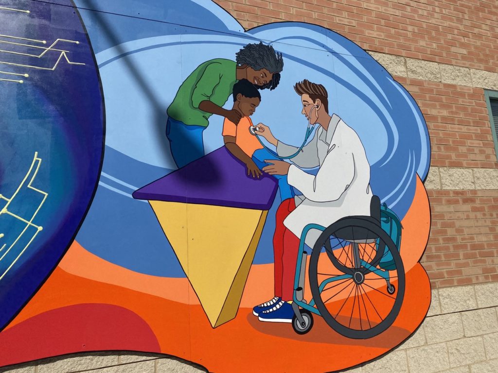 This is the final scene of the third stretch of the mural. It has a rounded shape as a frame, and the floor area of the scene is orange and red while the wall area of the scene is blue and light blue. The first person in the scene is a woman on the left. She has black skin, black, shoulder length hair styled in curls or dreadlocks, a green long sleeve shirt, and blue jeans. She is bending over to the right, with her right hand on the shoulder of her child. Her child is a young boy with black skin and short black hair. He is sitting on a counter with a yellow triangular base and a purple countertop. His legs are dangling off the right side of the counter. He wears an orange shirt and blue jeans. A doctor to his right is listening to his heartbeat with a stethoscope. The doctor is seated in a wheelchair with black wheels and a blue base. The doctor has short brown hair with an undercut, and their hair is coiffed. Their left hand is on the patient’s leg while their right hand uses the stethoscope. They are wearing a white medical coat, red pants, and dark blue sneakers.