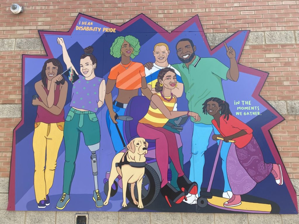 This is another section of the mural on another side of the building. It has a flat bottom, but becomes more jagged along the sides and especially the top. It is a group of people smiling out at the viewer. It has an indigo background with different shades of dark purple and blue. The person on the far left has brown skin, shoulder length dark brown hair, and is smiling. She is crossing her arms. Her left arm is holding an iPhone, and she rests her right elbow on top of her left arm. In her right hand, she holds a small disability pride flag. She wears a dark pink tank top, yellow corduroy pants, and purple shoes. To the right of her is a woman with white skin and brown hair tied in a bun on top of her head. She is raising her right arm in the air and making a fist. She is smiling. She is wearing a purple tank top with green polka dots, and teal pants. Her shoes are gray with yellow highlights. Her left leg is mechanical. To the right of her is a woman with brown skin, gold hoop earrings, and dark brown curly hair tied back in a bun. She is wearing a purple necklace, a yellow tank top with orange stripes, purple pants, and black high heels with red bottoms. She sits in a purple motorized wheelchair. On the ground to her left is a yellow labrador retriever serving as a guide dog, with a harness and a leash. The leash is held by a woman standing behind the scooter. She has black skin, green curly hair, an orange striped crop top, blue jeans, and a black belt. She is also wearing gold hoop earrings and a silver necklace. To the right of her, and still behind the woman in the motorized chair, is a man with white skin and short blond hair. He has blue eyes and is smiling. He is wearing a t-shirt that is white at the top, has a blue stripe in the middle, and a yellow stripe below that. He is wearing teal pants. Around his shoulders is the arm of the man to the right of him. This man has black skin, black hair, a black beard, and a collared, green polo shirt. He is smiling, and with his other hand he is holding up his pointer finger. He is wearing blue jeans and white shoes. In front of him is a young girl with black skin and brown and black hair in dreadlocks, tied in ponytails. He wears a red long sleeved shirt, a long purple paisley skirt, and pink sneakers with white bottoms. She looks to be entering the scene on a scooter that has a yellow platform and handle but a purple pole.